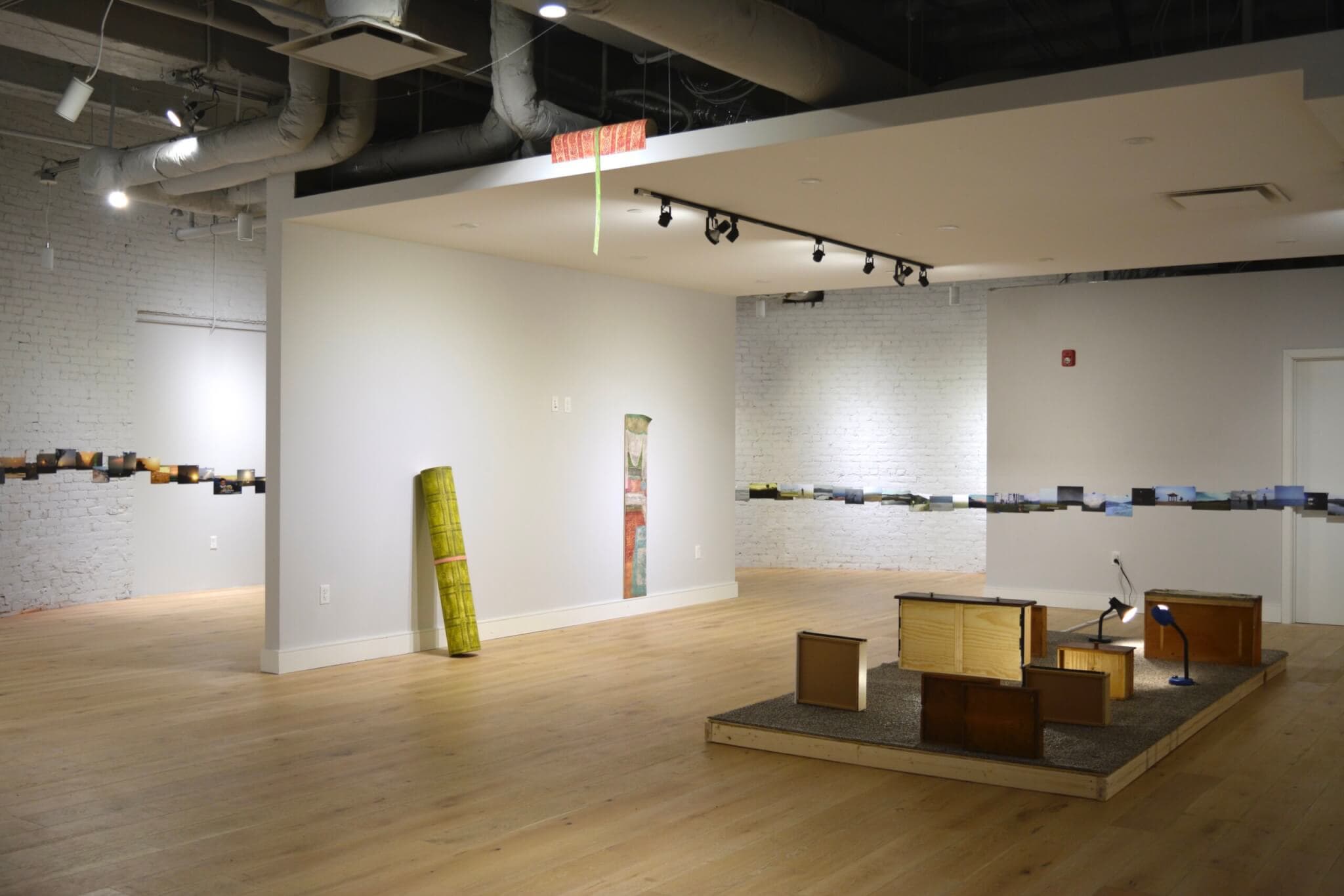 Photographs line the perimeter of a gallery space that features wooden sculptures and dresser drawers turned on their sides.
